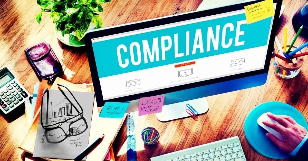 A desktop monitor displays the word “COMPLIANCE,” highlighting website legal compliance. The desk has a keyboard, notes, papers, a plant, calculator, coffee, and a person using a mouse.