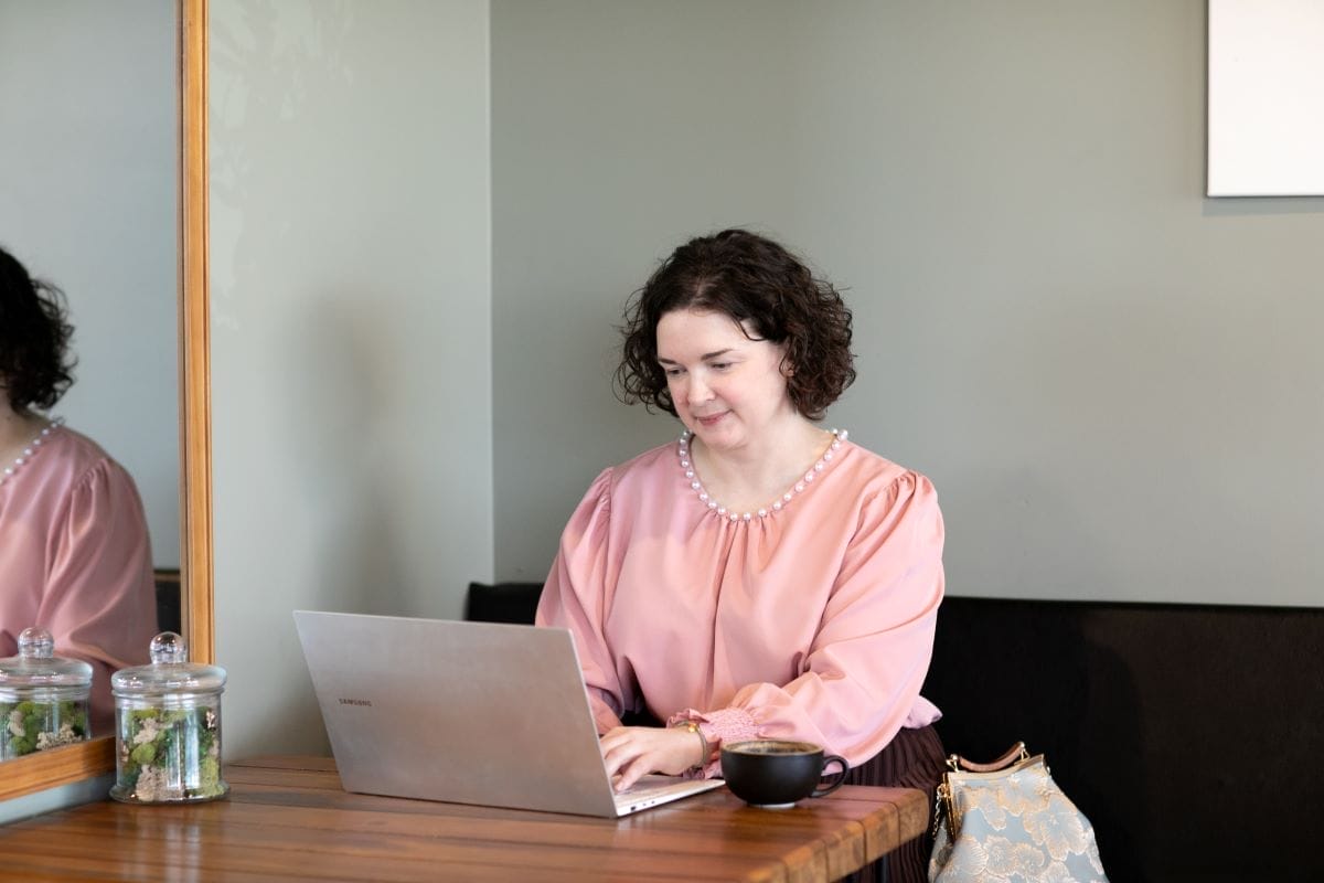 A woman in a pink blouse sits at a wooden table, focused on copywriting and content writing on her laptop. A coffee cup, purse, and glass jars are arranged neatly beside her.
