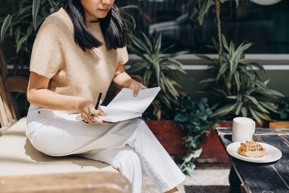 A woman sits outdoors on a bench, holding a pen and an open notebook for copywriting. Nearby, a table holds a cup of coffee and a pastry, surrounded by lush plants in the background.