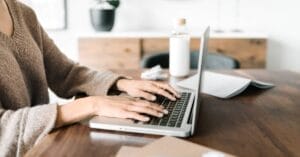 Person typing on a laptop at a wooden table with a notebook, bottle, and blurred background items visible—perfect for those seeking high quality stock photos.