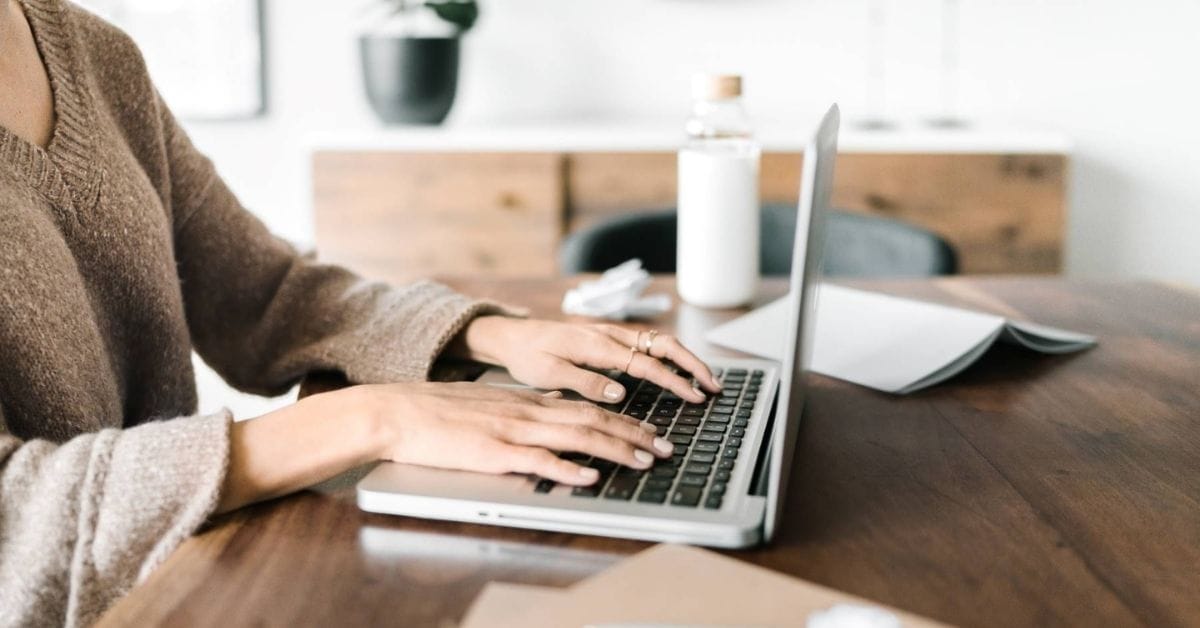 Person typing on a laptop at a wooden table with a notebook, bottle, and blurred background items visible—perfect for those seeking high quality stock photos.