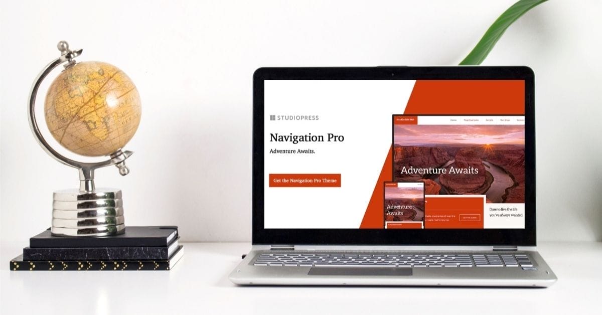 A laptop displaying the StudioPress Navigation Pro theme on the Genesis Framework sits on a white desk next to a globe and two stacked books, with a green plant partially visible.