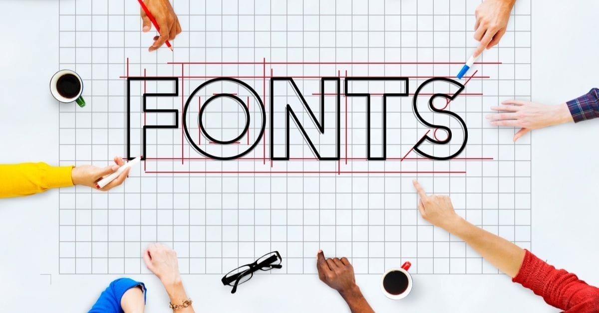 Several hands point to and work on the word "FONTS" outlined on a large grid paper, surrounded by coffee cups, glasses, and office supplies—perfect for brainstorming font pairings for websites.