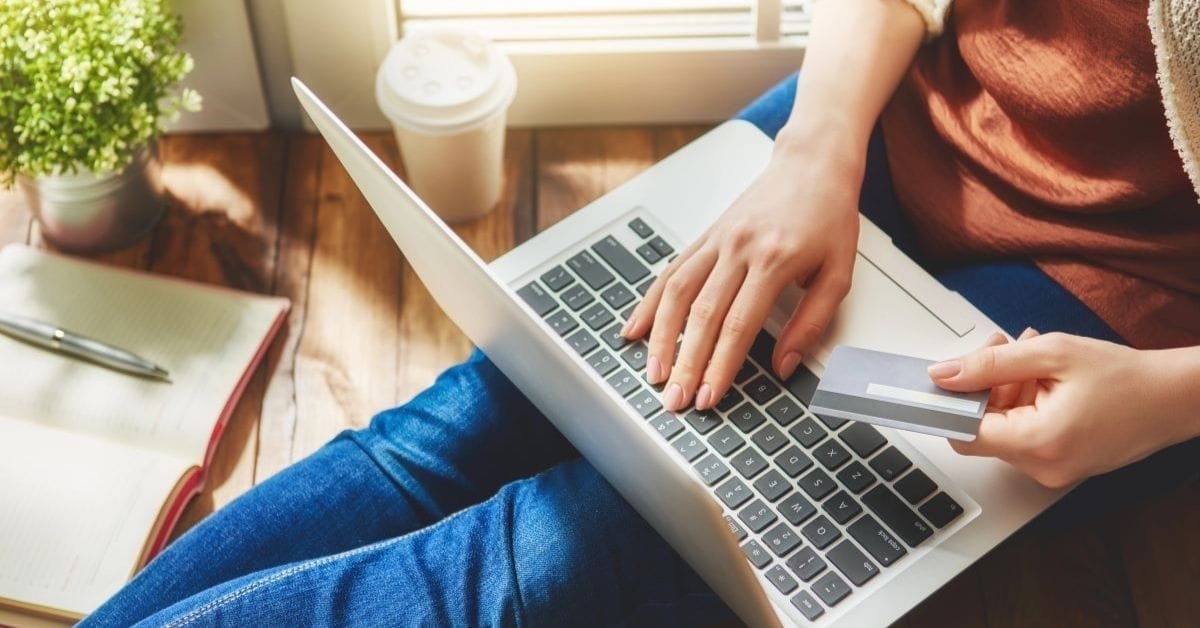 Person sitting on the floor using a laptop, holding a credit card—ready to buy SendOwl digital products. A notebook, plant, and coffee cup are nearby, creating a cozy workspace.