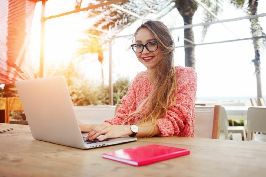 A woman with glasses and long hair works on a laptop at an outdoor table with palm trees in the background. A red notebook rests on the table beside her, hinting at her dedication to her online business.