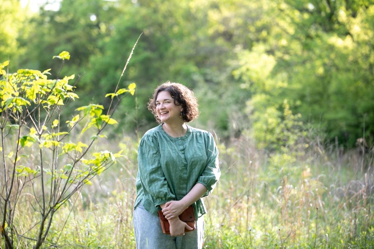 A woman with curly hair and glasses stands outdoors in a sunlit field, wearing a green shirt and smiling while holding a brown purse, embodying the creativity found in branding and web design.