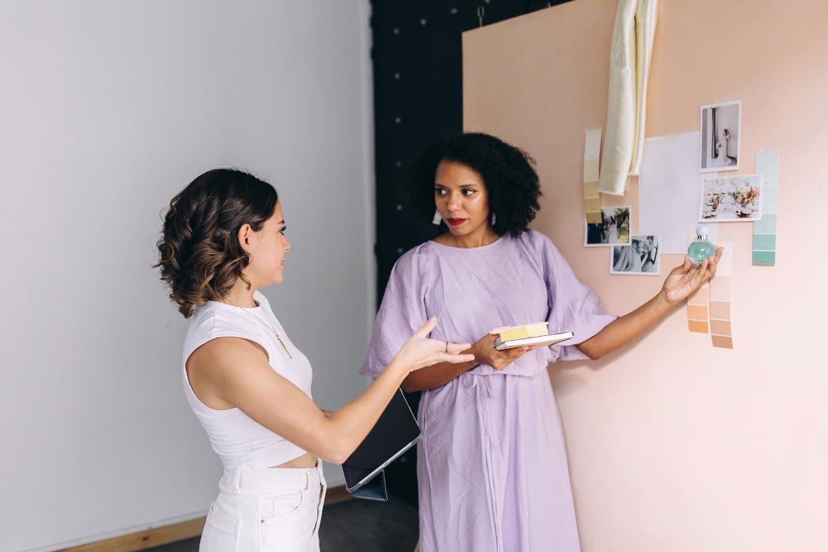 Two women stand by a peach-colored wall with color swatches and photos, discussing interior design ideas and how branding and web design can tie the look together. One holds notebooks while the other gestures towards the wall.