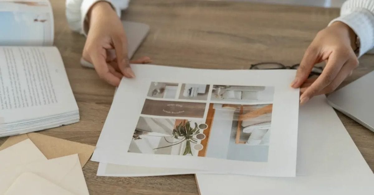 Person holding a printed sheet with interior design photos on a table, surrounded by a book, envelopes, and a laptop—perfect for brainstorming branding and web design inspiration.