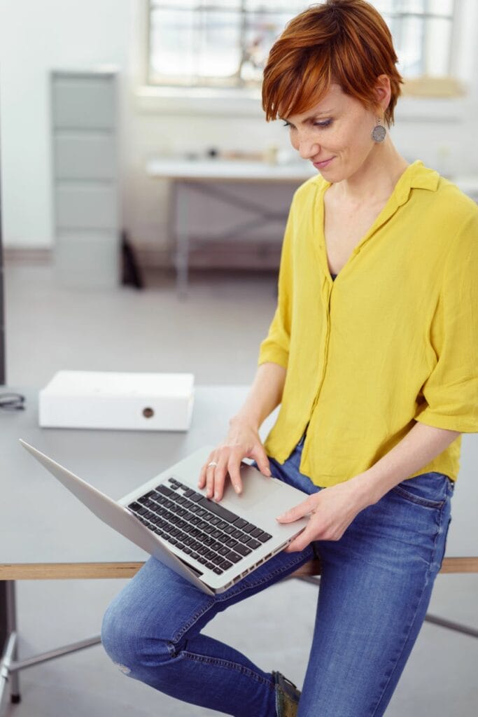 A person with short hair, wearing a yellow blouse and jeans, sits on a desk using a laptop in a bright, minimalist office setting, immersed in conducting an SEO website audit.