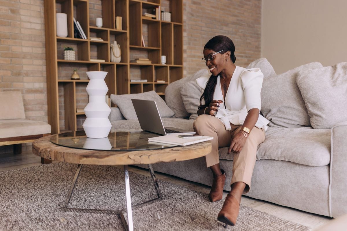A woman sits on a beige sofa, looking at an open laptop on a wooden coffee table in a modern living room, preparing to audit my website with shelves and decor in the background.