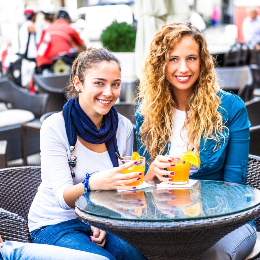 Two women seated at an outdoor cafe table, smiling, each holding a glass of orange juice garnished with an orange slice, as they discuss the latest website wellness review.