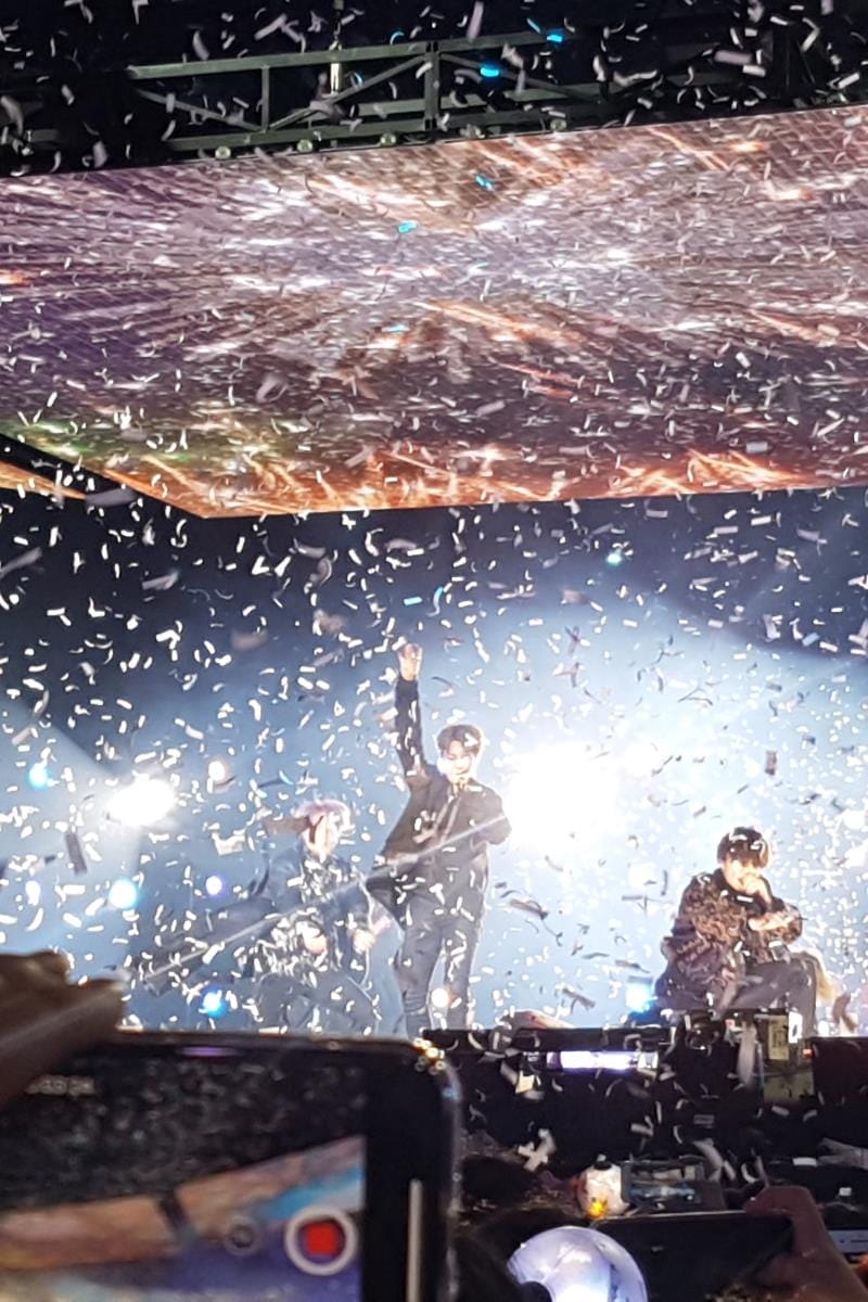 Performers on stage surrounded by confetti, with one holding up a hand. Bright lights and a large screen are visible in the background.