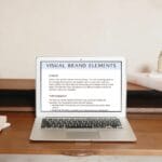 A laptop displaying a presentation titled "Visual Brand Elements" is on a wooden desk with notebooks and decorative items around it.