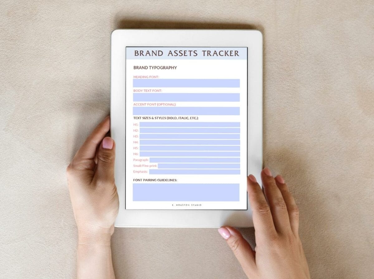 Person holding a tablet displaying a brand assets tracker document, including brand typography and text sizes, on a neutral surface.