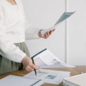 Person in a white sweater standing at a table, holding papers and a pen, with notebooks and documents spread out in front.