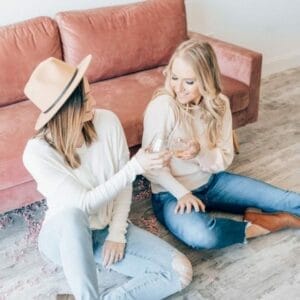 Two women sitting on the floor and clinking glasses in front of a pink sofa. One wears a hat, both are smiling.