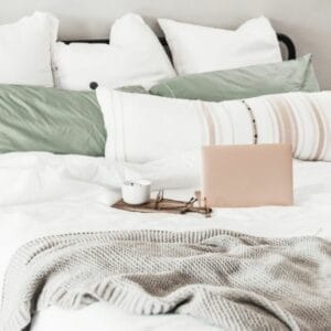 A cozy bed with white and green pillows, a beige blanket, and a small brown tray holding a closed pink laptop and glasses.