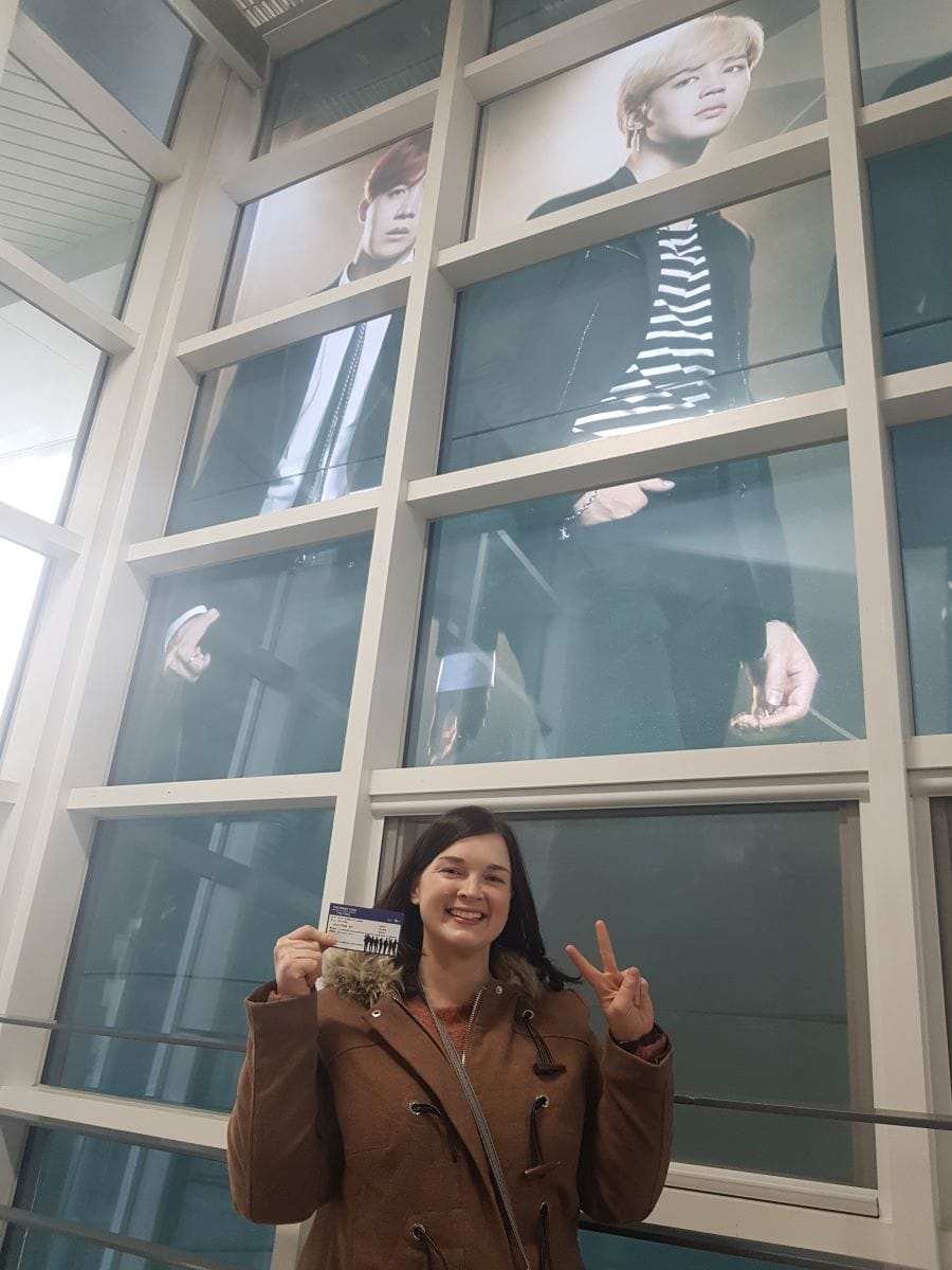 A person smiling and holding a booklet in front of large posters of two individuals inside a building.