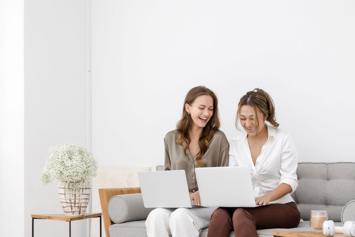Two women sit on a gray sofa working on laptops, smiling. A small table with a vase of flowers and a coffee cup is next to them in a bright, minimalist room.