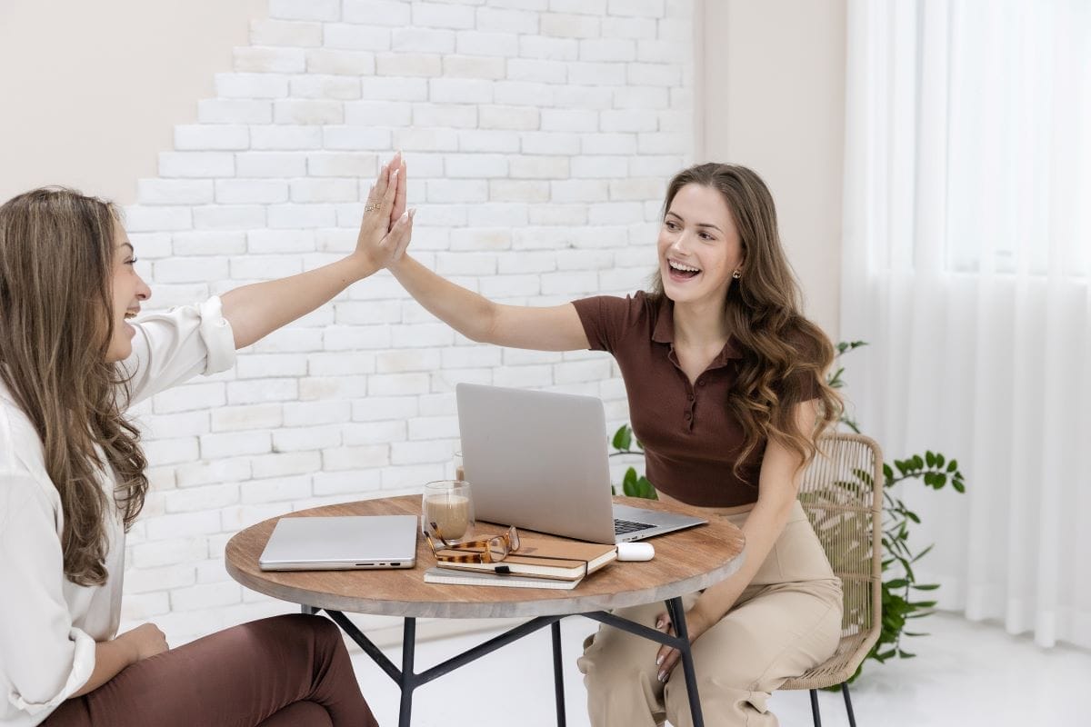 Two women sitting at a round table with laptops, high-fiving and smiling in a bright room with white walls and plants.