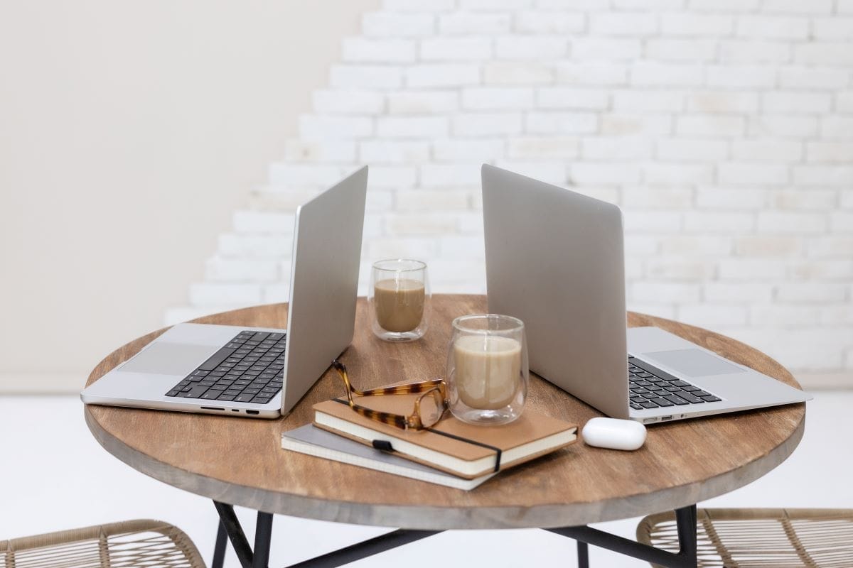 Two open laptops, two glasses of coffee, notebooks, eyeglasses, and wireless earbuds on a round wooden table against a white brick wall.