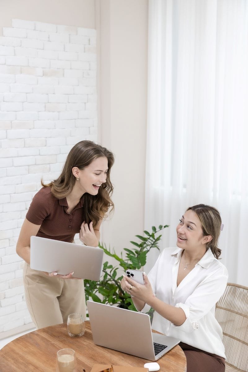 Two women smiling and talking in an office setting, each holding a laptop; one is seated at a desk while the other stands beside her.