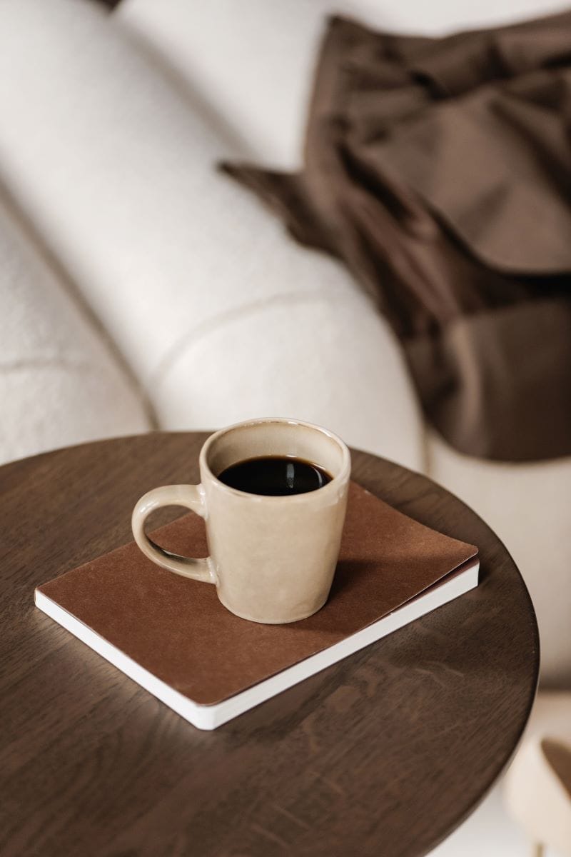 A beige ceramic mug filled with black coffee sits on a closed brown notebook atop a round wooden table, with a cream-colored sofa and brown blanket in the background.