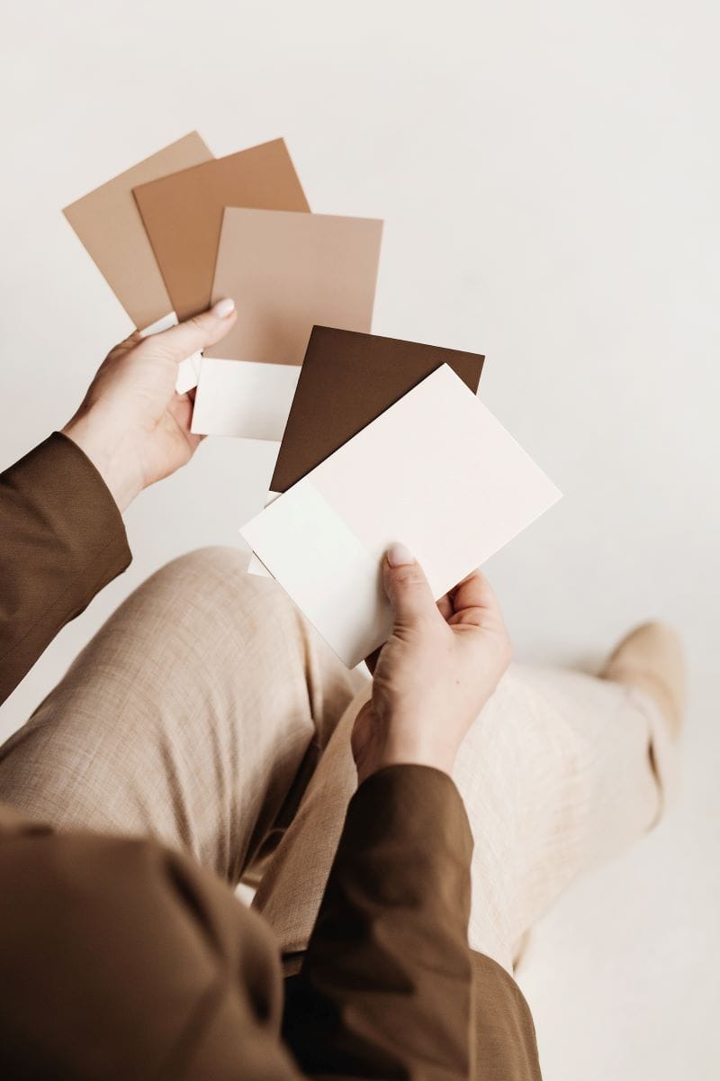 A person sits holding several paint color swatches in various shades of brown and beige against a neutral background.