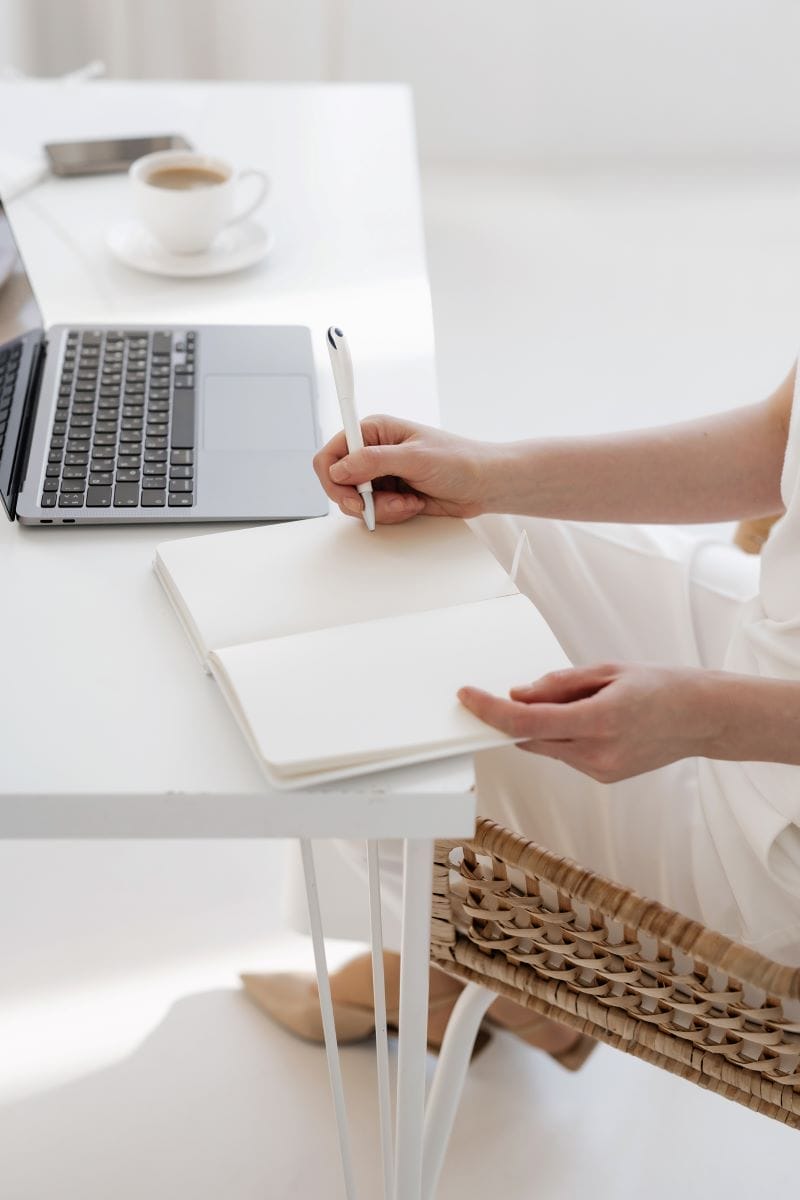 A person sits at a desk with a laptop and coffee, writing in a blank notebook with a pen.