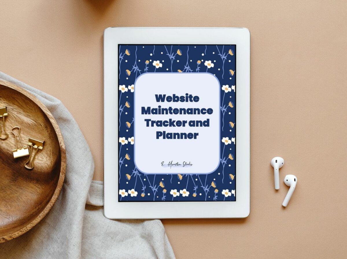 A tablet displays a "Website Maintenance Tracker and Planner" cover with a blue floral design. Nearby are wireless earbuds, a wooden tray, and gold binder clips on a beige surface.