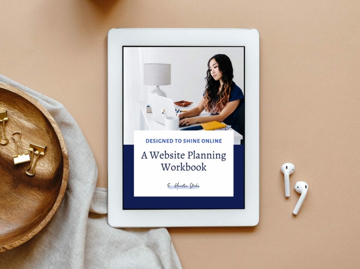 An iPad displays the cover of "A Website Planning Workbook" on a beige desk with a wooden tray, paper clips, fabric, and wireless earphones nearby.