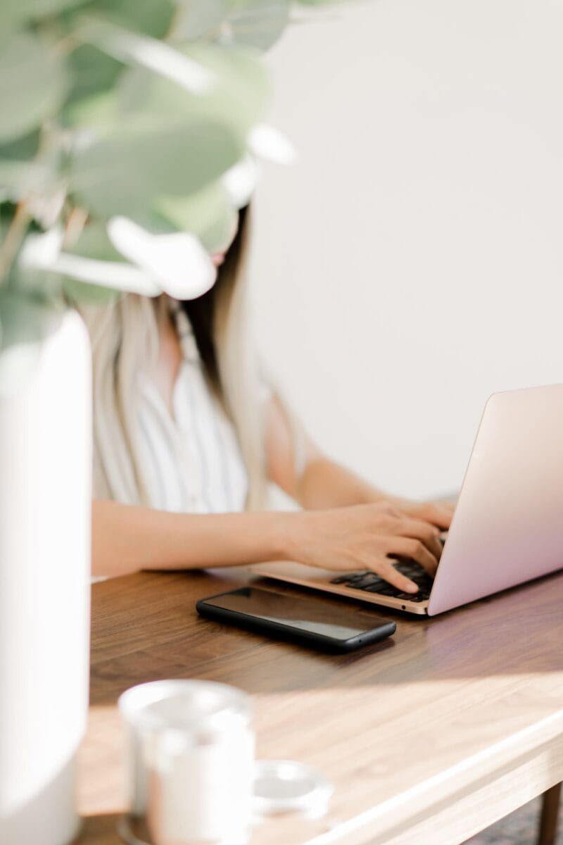 Person typing on a laptop at a wooden table with a smartphone nearby. A plant is in the foreground.