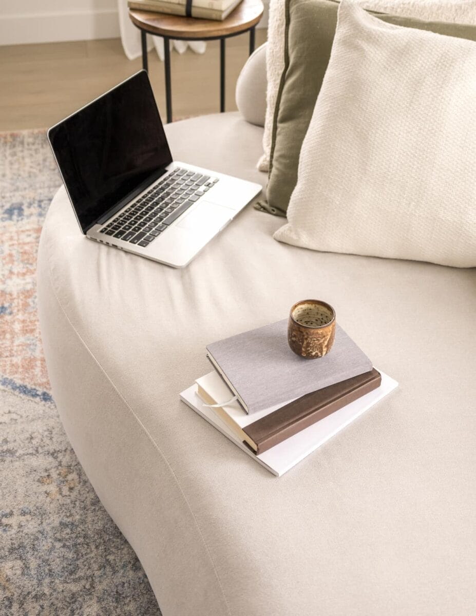 Open laptop on a beige sofa with a stack of books and a coffee mug nearby. Decorative cushions are in the background.
