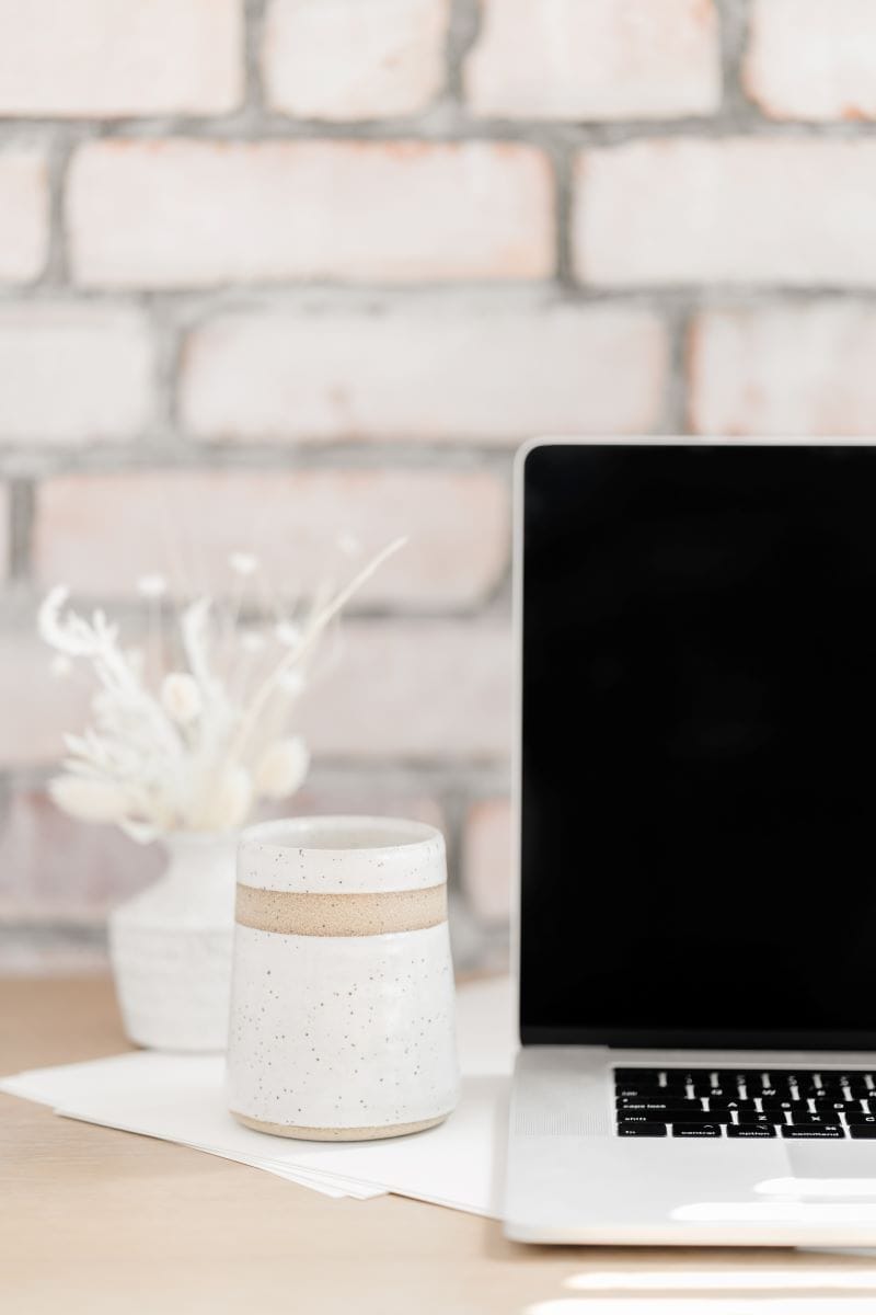A ceramic cup sits on a desk next to an open laptop, with a small vase of dried flowers in the background against a white brick wall.