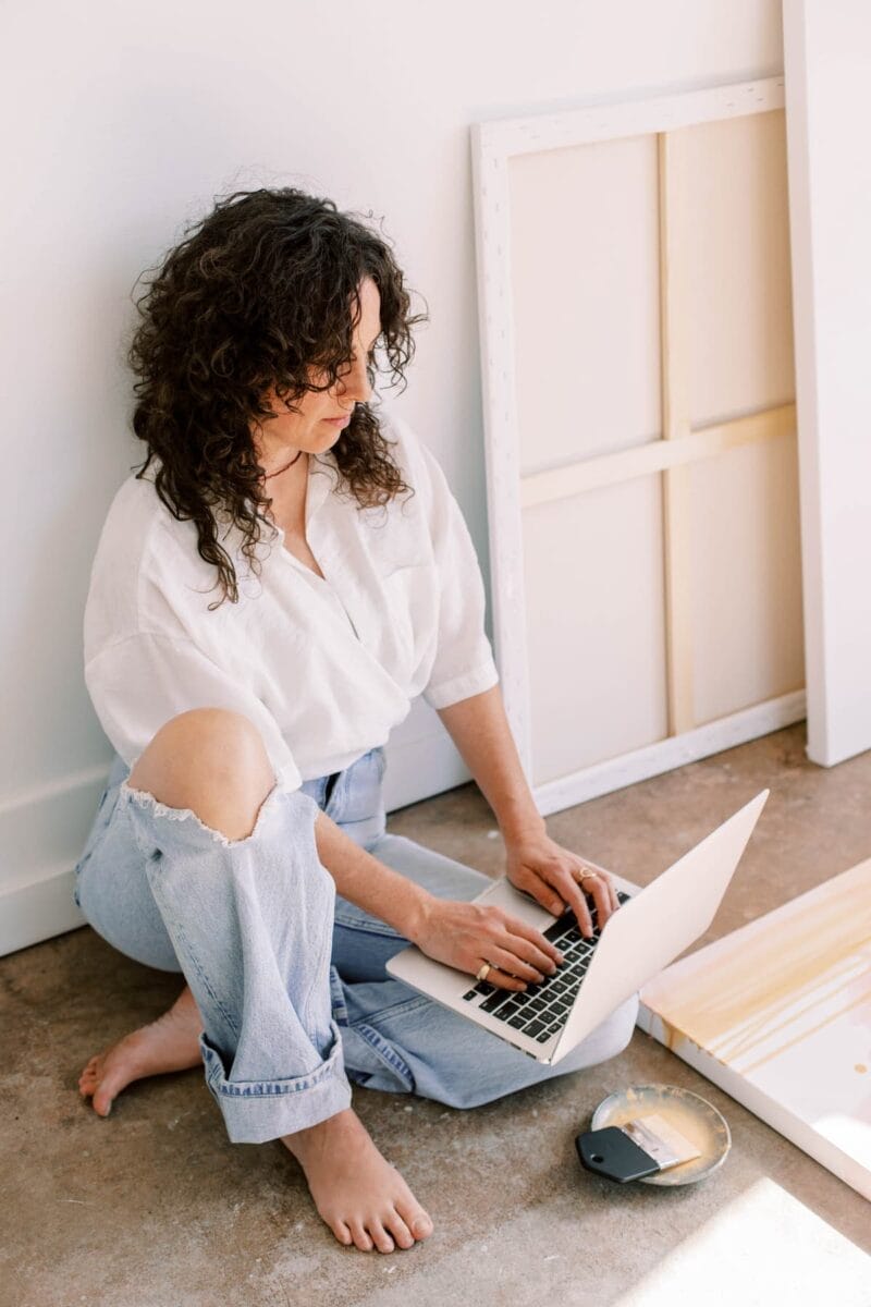 Person with curly hair sitting on the floor, using a laptop. They are wearing a white shirt and ripped jeans. Art supplies and a canvas are nearby.