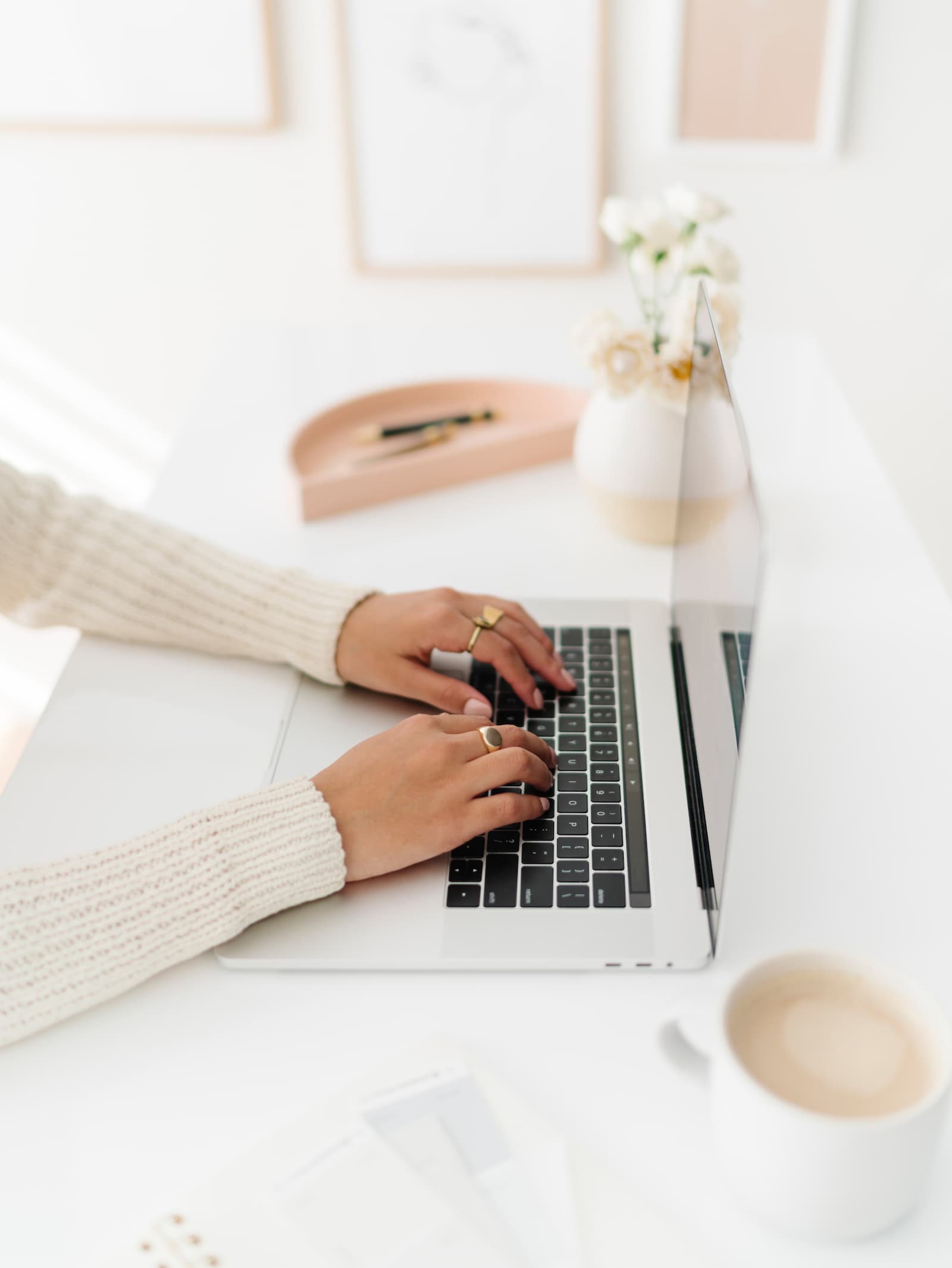 Person typing on a laptop at a white desk with a cup of coffee, a notebook, and a vase of flowers.