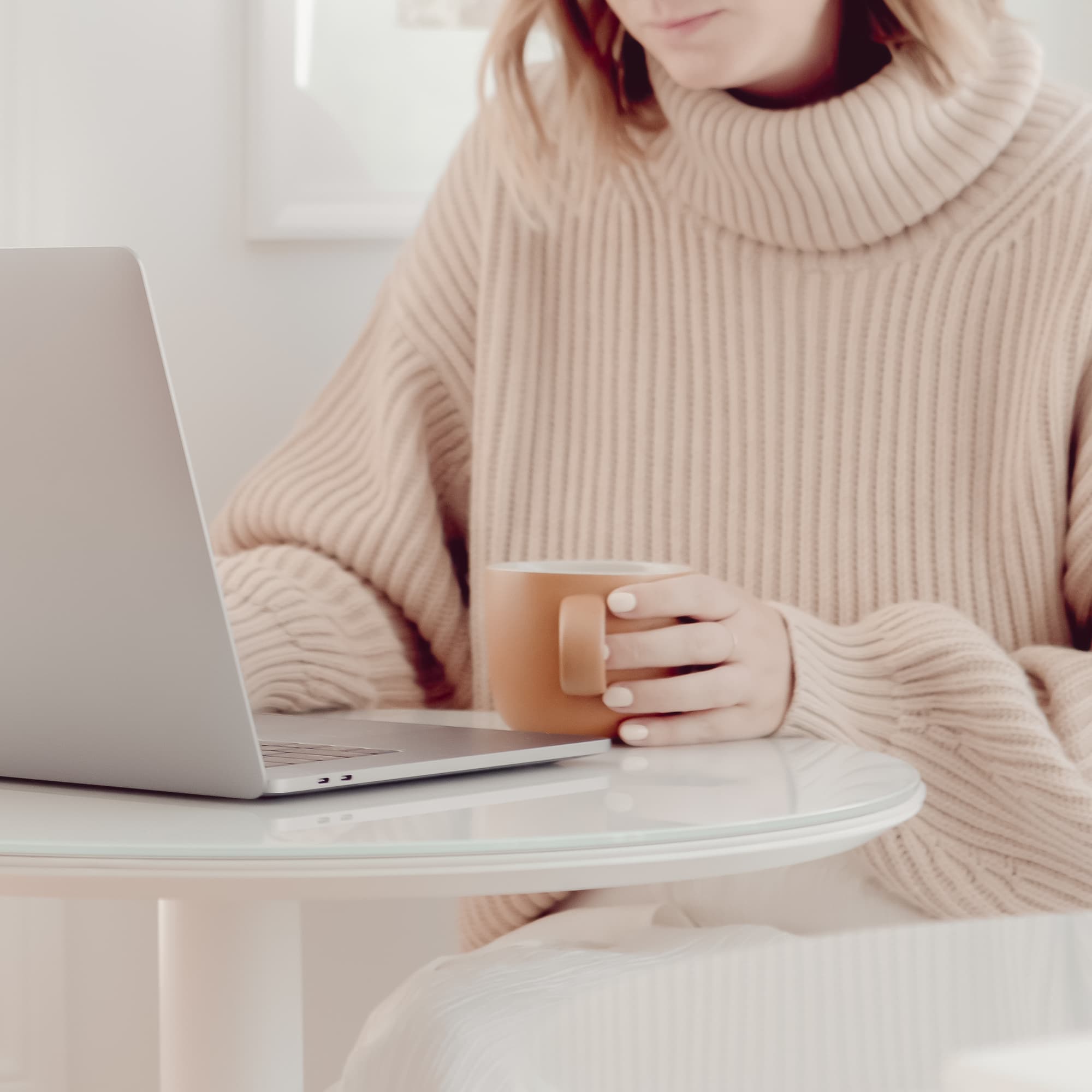Person in a beige sweater holding a mug, sitting at a round table with an open laptop.
