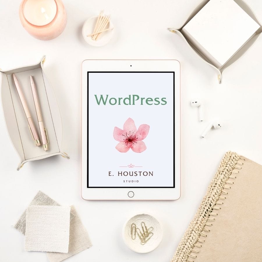Tablet displaying a WordPress cover with a pink flower, surrounded by office supplies, a candle, AirPods, and a woven notebook on a white desk—perfect inspiration for your 30 day website challenge.