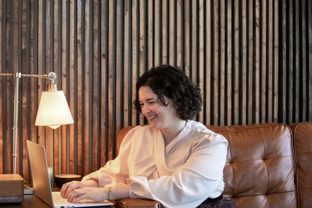 A woman sits on a brown leather couch, smiling while performing a website audit on her laptop, with a lamp and wooden slat wall in the background.