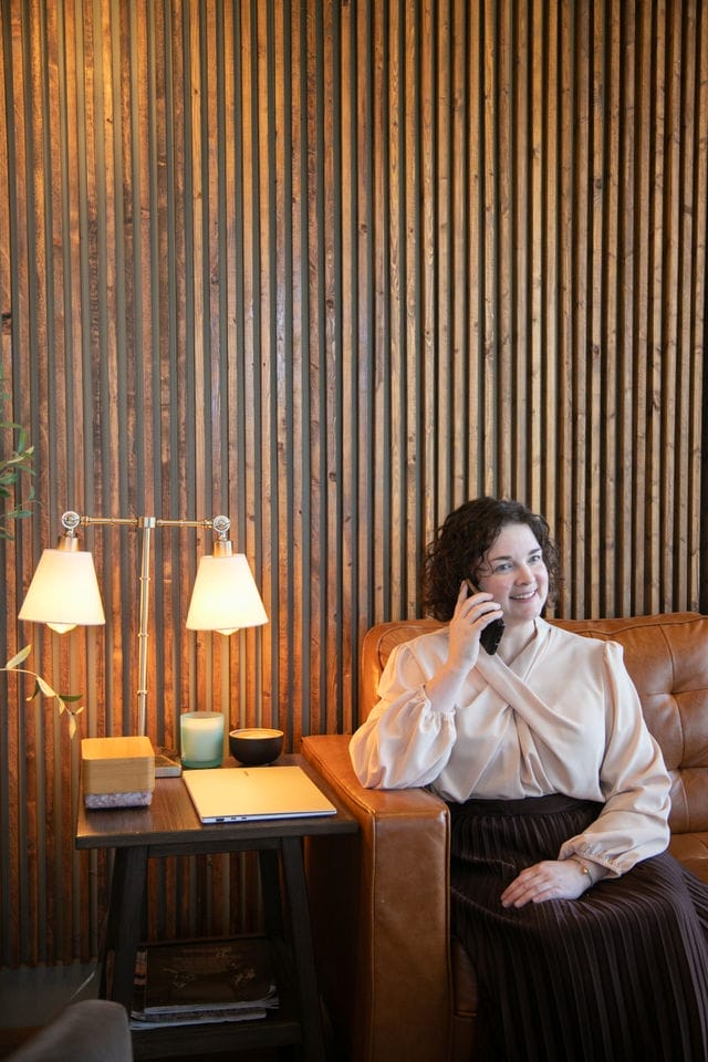Woman sitting on a brown leather couch, smiling while talking on the phone—perhaps arranging a website design contact—beside a side table with two lit lamps, books, and a cup, in a room with a wooden slat wall.