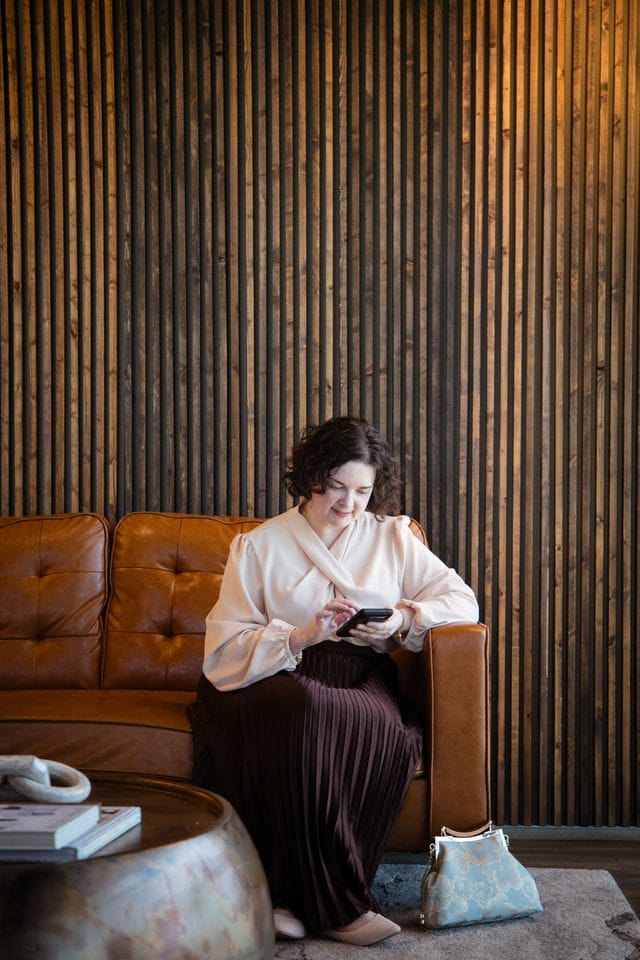 A woman sits on a brown leather sofa, looking at a smartphone—perhaps reviewing a website audit. A blue handbag rests beside her, and a round coffee table with books is in front of her. The wall behind features vertical wooden slats.