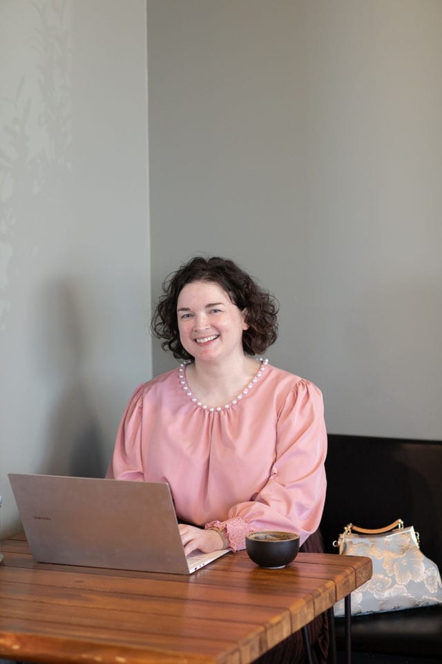 A woman in a pink blouse sits at a wooden table working on a laptop, focusing on website success, with a cup and a purse beside her.