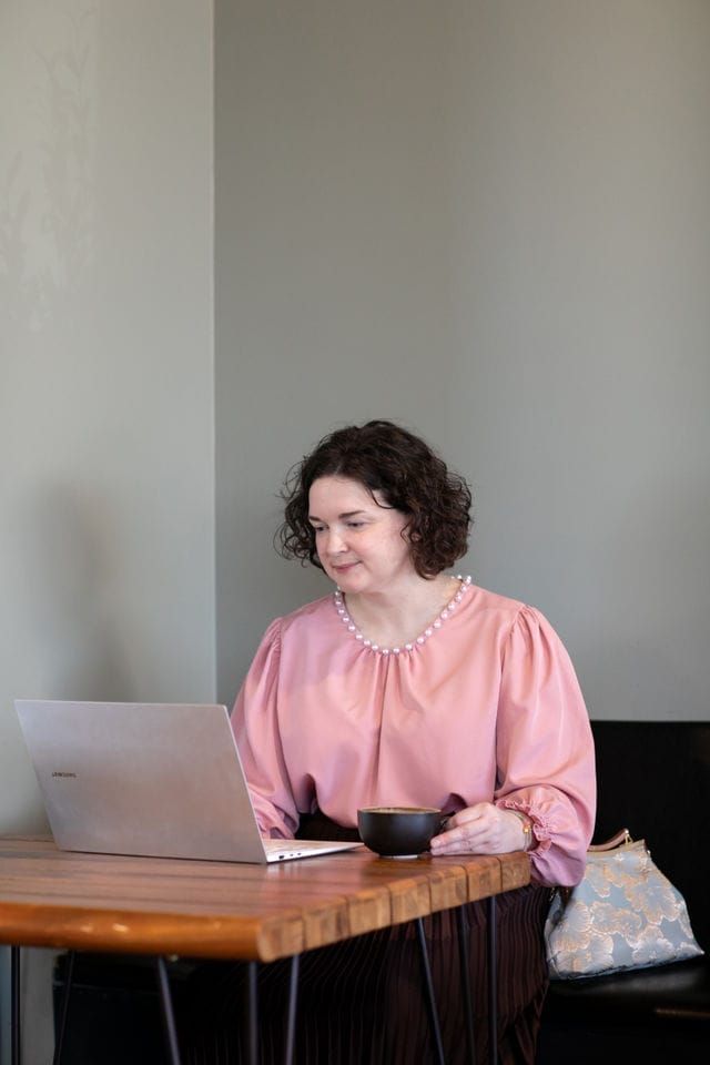 Woman with curly hair in a pink blouse sits at a wooden table, using a laptop and holding a black cup, focused on website growth, with a light-colored bag beside her.