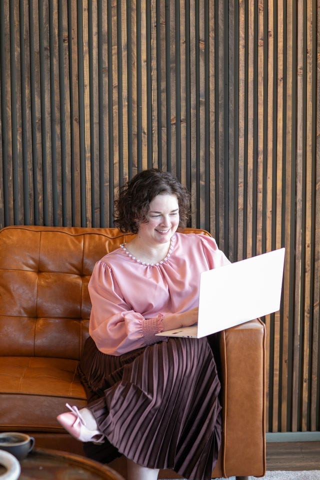 A woman sits on a brown leather couch, smiling as she works on a laptop. She is wearing a pink blouse and pleated skirt, focused on creating a professional website makeover.