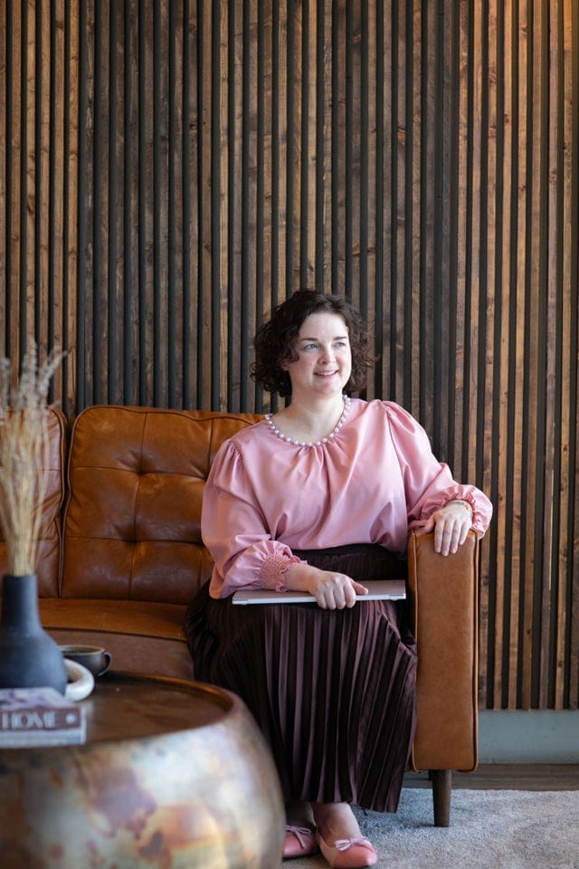 A woman with curly hair sits on a brown leather sofa, wearing a pink blouse and pleated skirt, holding a tablet as she reads about WordPress safety, with a striped wooden wall in the background.