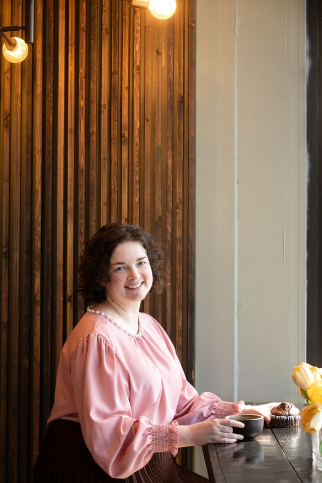 A woman with curly hair wearing a pink blouse sits at a wooden table by a window, enjoying coffee and a cupcake while working on keyword research for website optimization.
