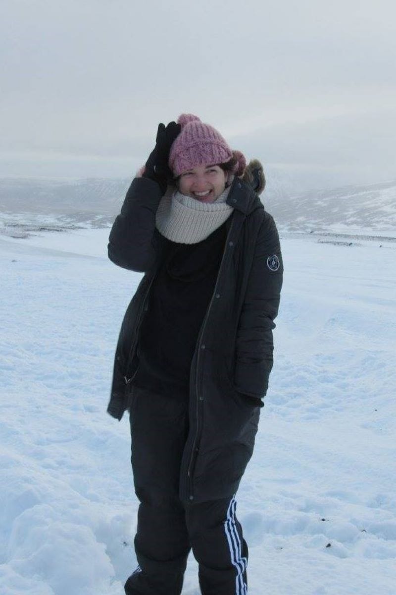 Person standing outdoors in a snowy landscape, wearing a pink knit hat, scarf, dark coat, and track pants, smiling at the camera—perfect inspiration for any WordPress and Showit Web Designer seeking winter-themed visuals.
