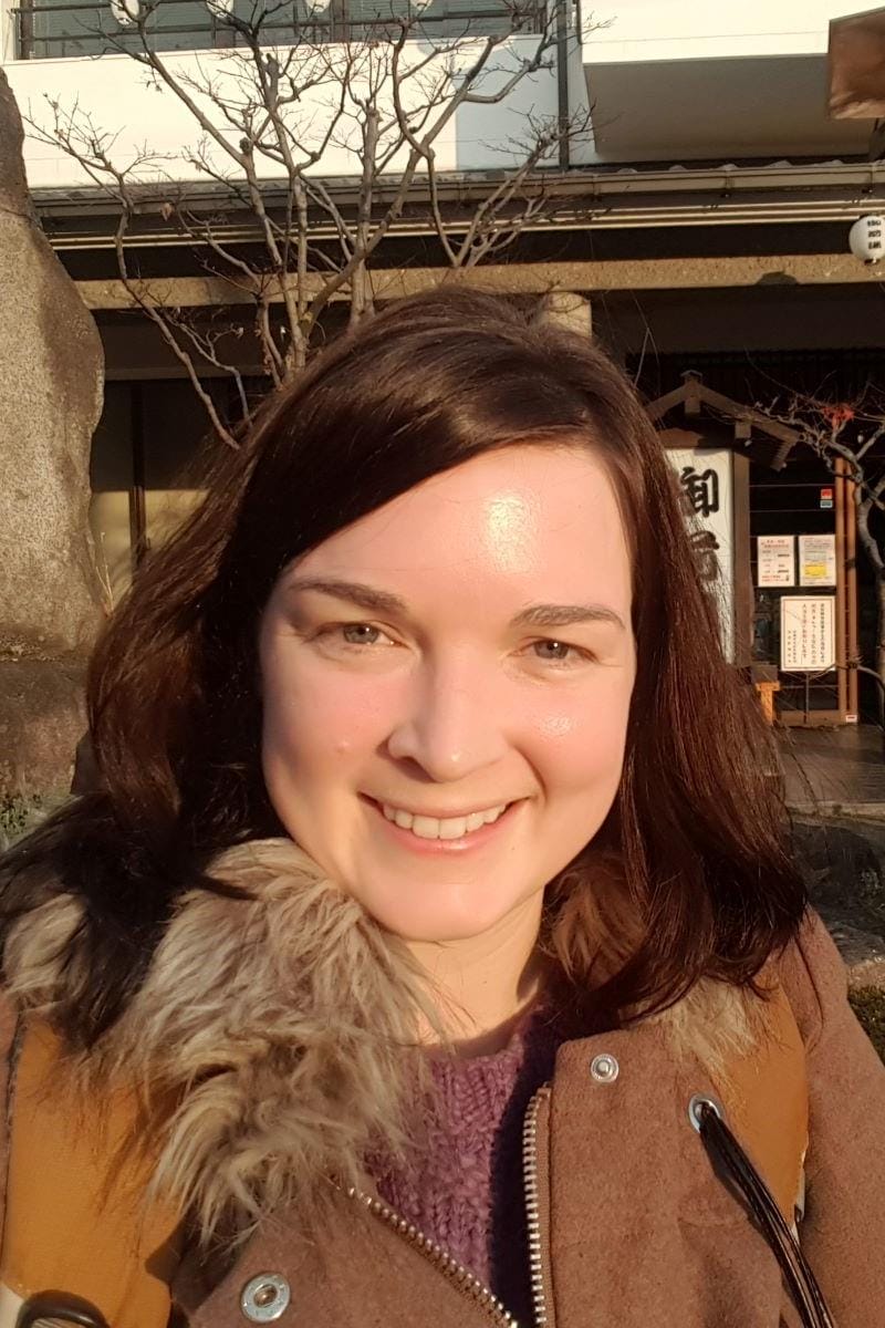 A woman with brown hair and a brown coat smiles outdoors in sunlight, with a building and leafless tree in the background, capturing the fresh energy of a website makeover.