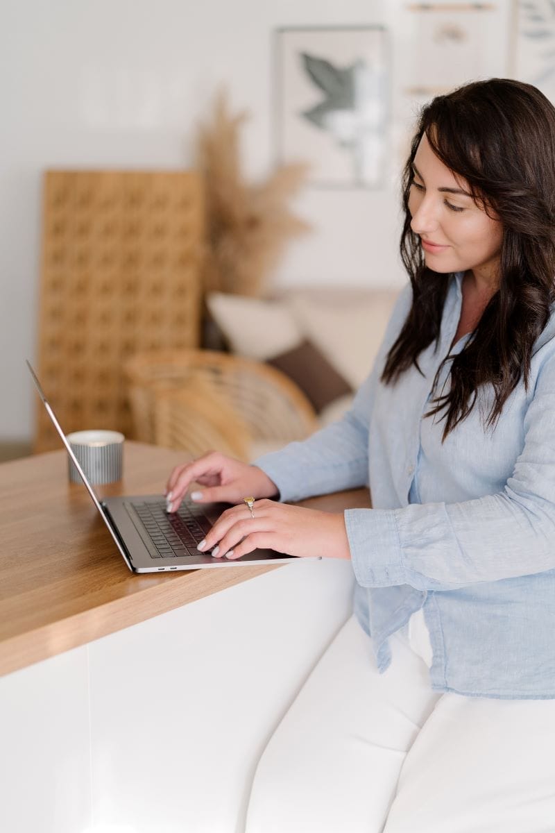 A woman sits at a wooden counter working on a laptop, with a mug nearby. She is dressed in a light blue shirt and white pants in a softly lit room.