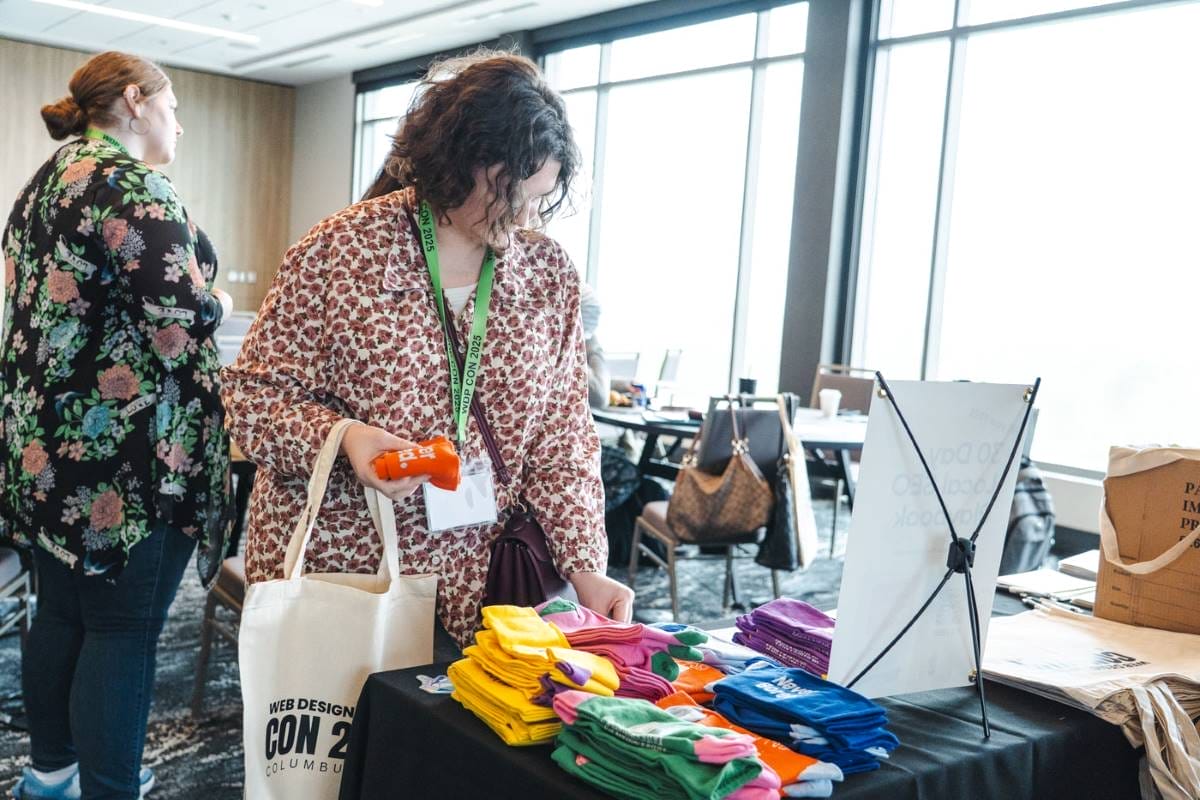 A person with a tote bag browses colorful folded t-shirts on a display table at an indoor event; other people and tables are visible in the background.